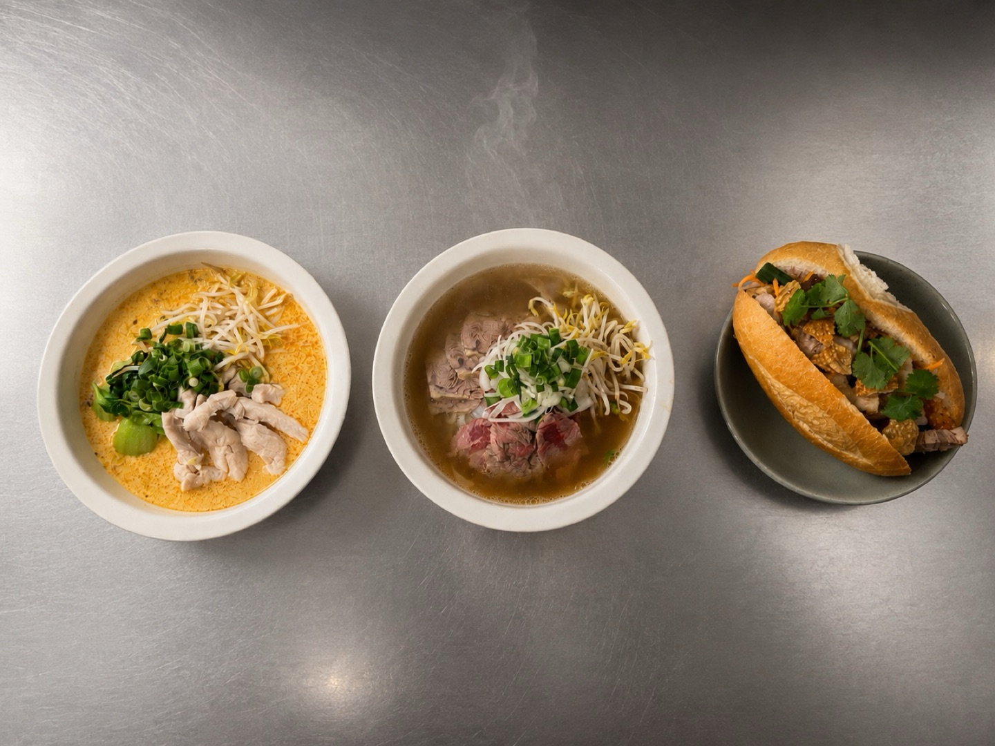 Two noodle soup bowls and a bánh mì lined up on the stainless steel bench