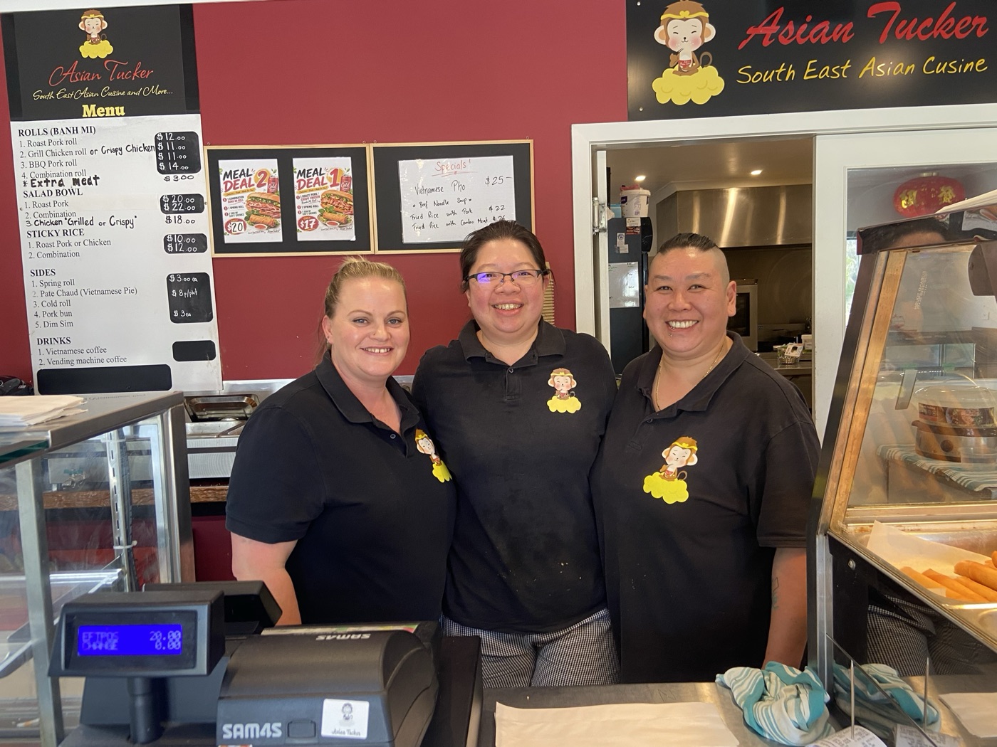 The Asian Tucker crew smiling behind the counter, menu board and monkey logo behind them
